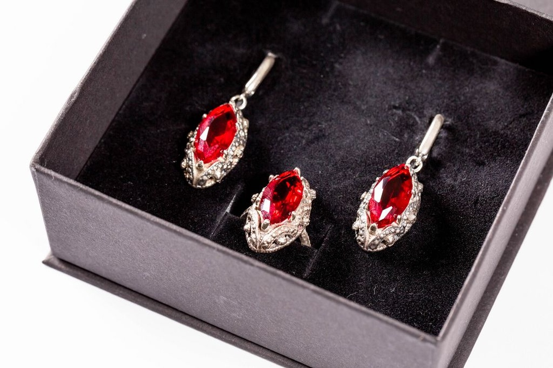 Ett smyckeskrin i svart sammet innehållande ett matchande set med röda ädelstenar, troligen rubiner eller granater. Setet består av örhängen och en ring, alla med marquise-slipade röda stenar omgivna av små diamanter eller kristaller i silverfärgade infattningar.